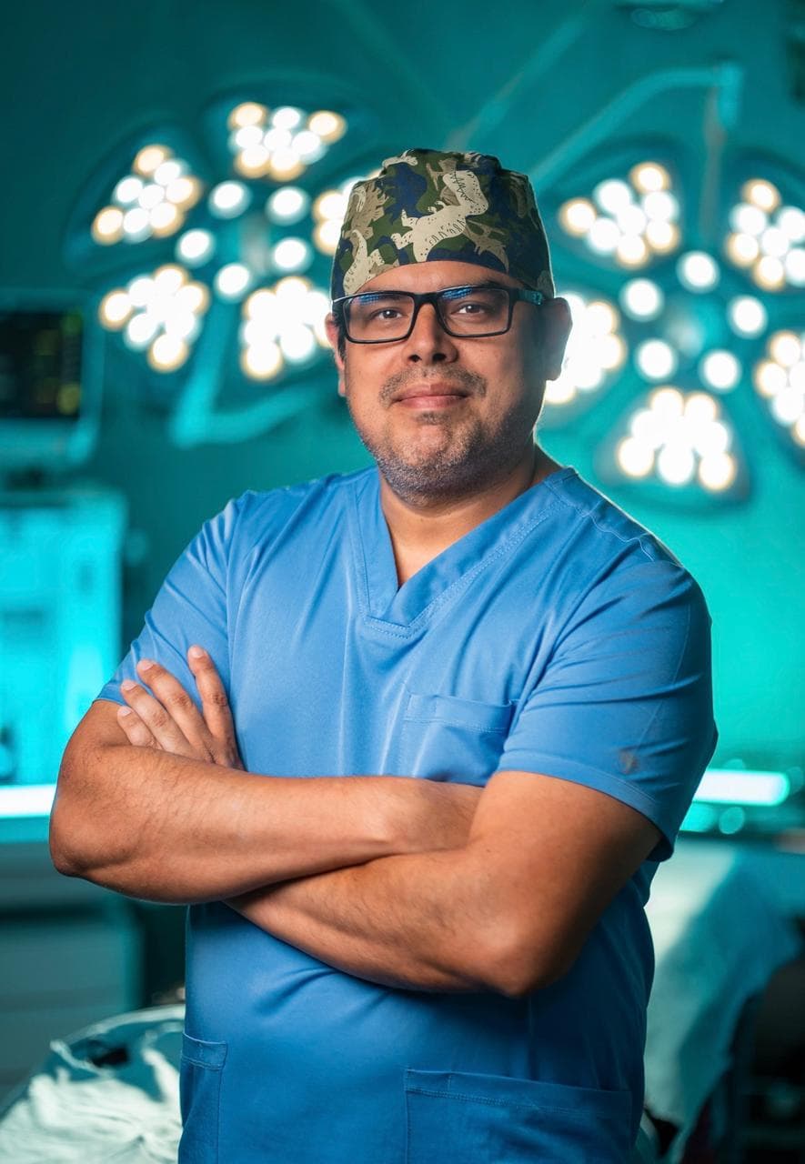 Dr. Mardonio Euscátigue con uniforme azul y brazos cruzados, frente a las luces del quirófano